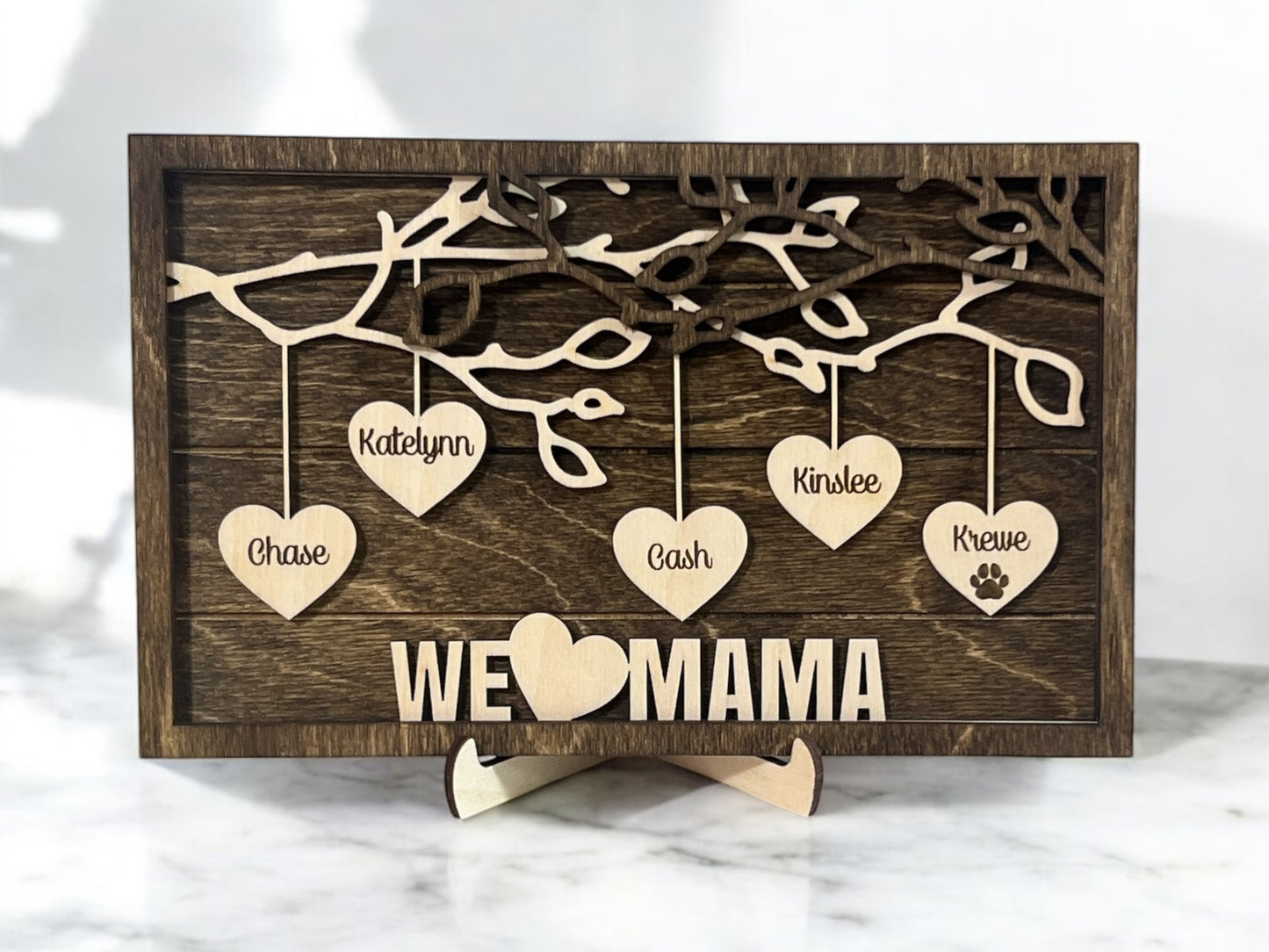 A wooden family hearts sign with multiple hanging hearts, each engraved with a name, and the phrase 'WE love MAMA' on a shiplap background with a dark walnut stained back.
