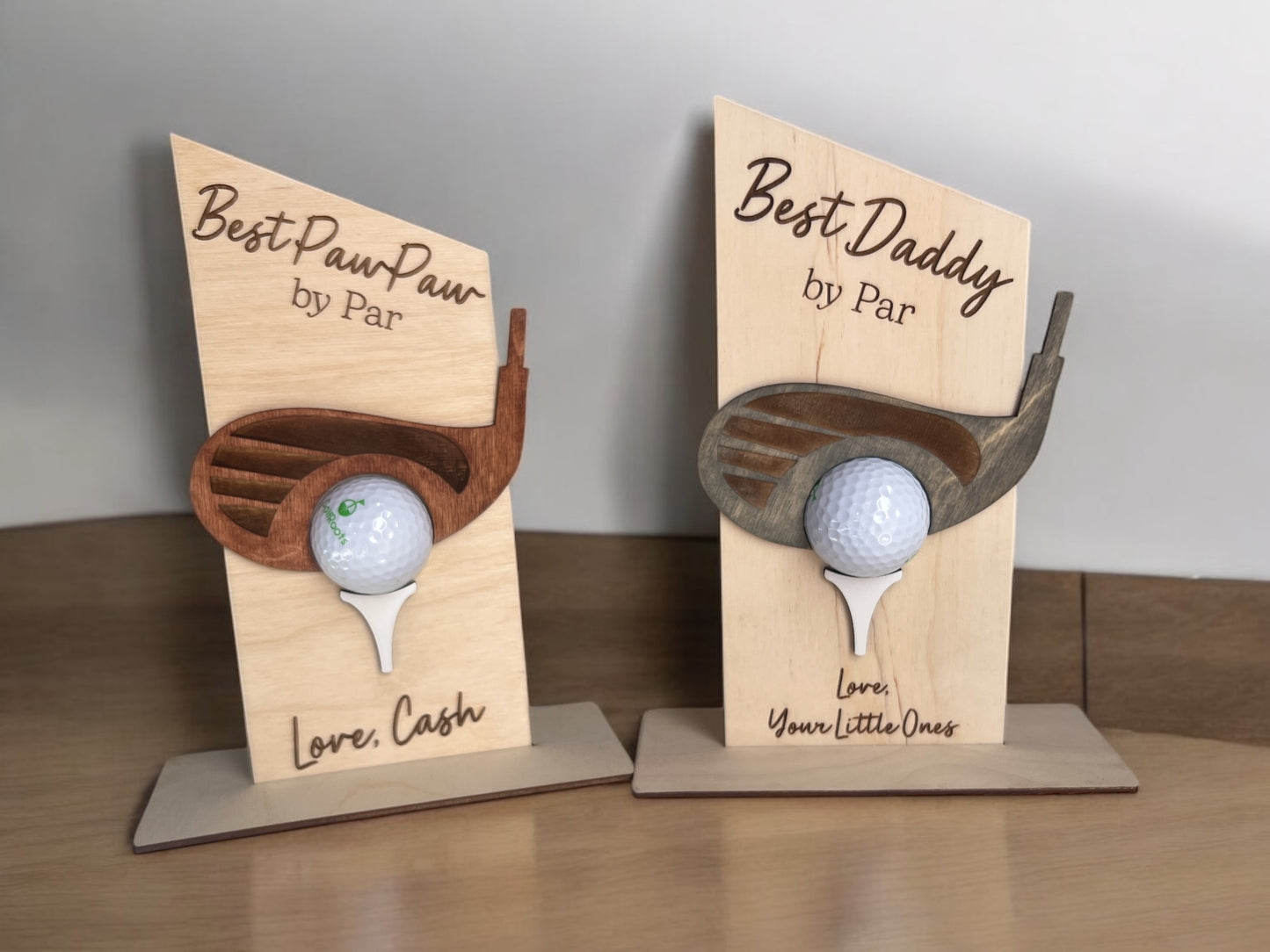 Two wooden golf ball holders with a title and personalized message printed on a wooden stand, each holding a golf ball. One is labeled 'Best PawPaw' and the other 'Best Daddy', both with a 'Love' message.