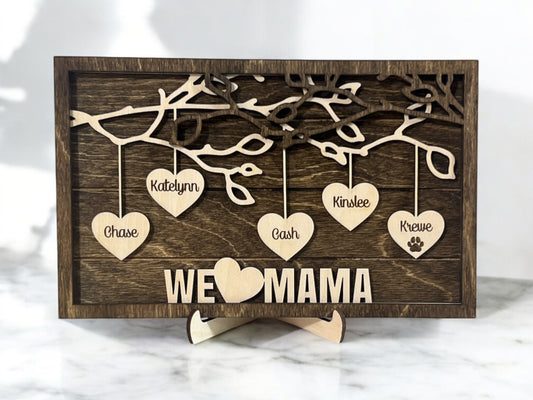 A wooden family hearts sign with multiple hanging hearts, each engraved with a name, and the phrase 'WE love MAMA' on a shiplap background with a dark walnut stained back.