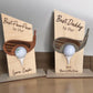 Two wooden golf ball holders with a title and personalized message printed on a wooden stand, each holding a golf ball. One is labeled 'Best PawPaw' and the other 'Best Daddy', both with a 'Love' message.