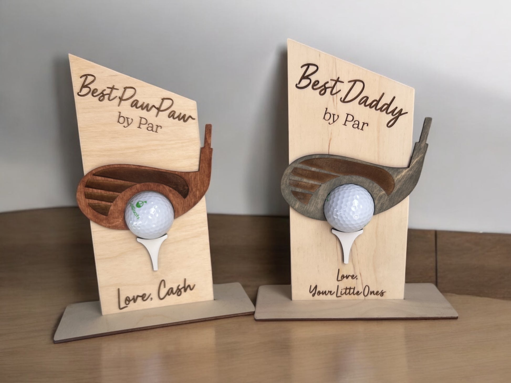 Two wooden golf ball holders with a title and personalized message printed on a wooden stand, each holding a golf ball. One is labeled 'Best PawPaw' and the other 'Best Daddy', both with a 'Love' message.