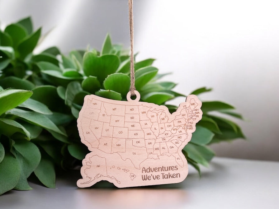 USA map ornament with 'Adventures We've Taken' text, hanging against a blurred green plant background.